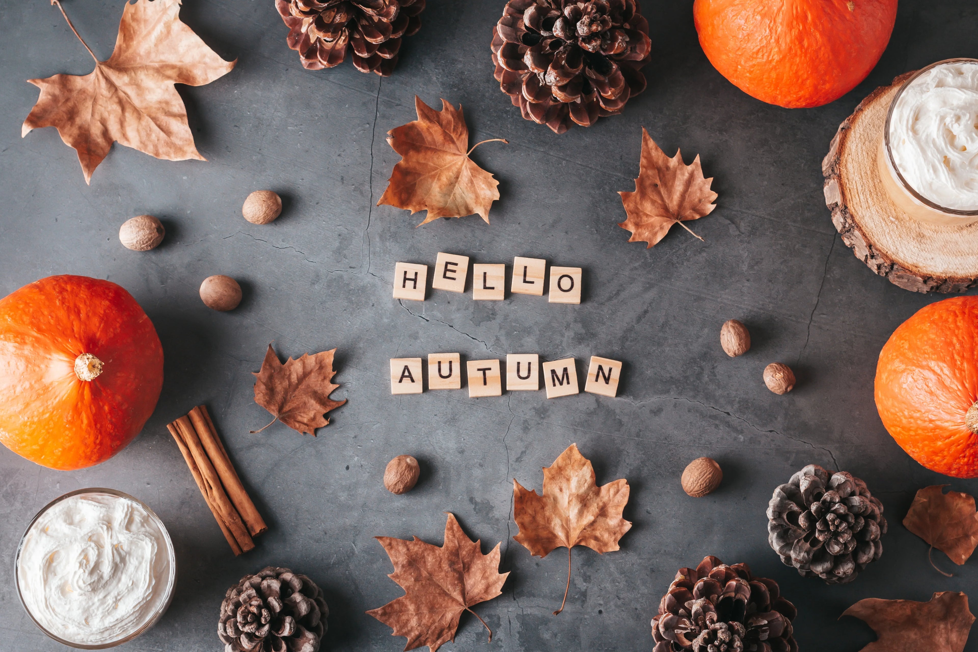 Décor automnal avec les lettres de Scrabble qui épellent "Hello Autumn", des feuilles mortes, des citrouilles, des pommes de pin et des bâtons de cannelle sur un fond noir style ardoise.